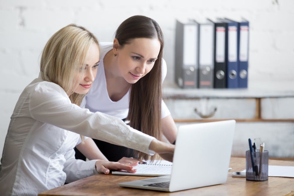 Eine Frau sitzt vor ihrem Laptop, zeigt auf den Bildschirm, während eine andere Frau daneben steht und auf den Bildschirm schaut.