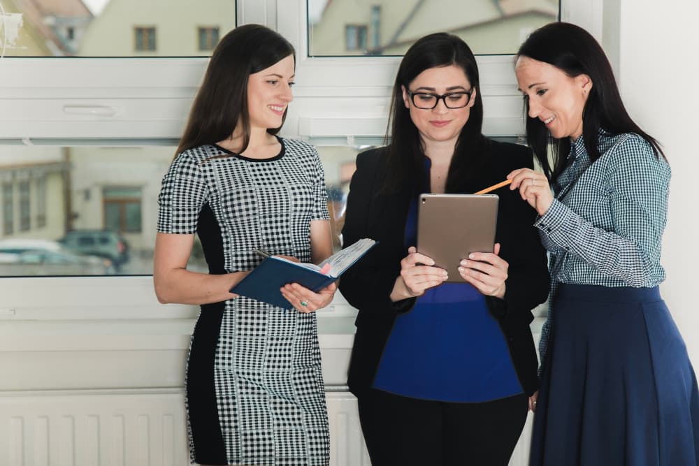 Drei Geschäftsfrauen betrachten ein Tablet während eine von ihnen ein Buch hält.