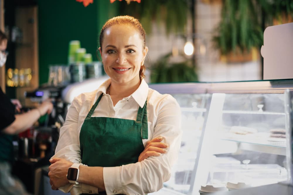 Eine Frau in grüner Schürze steht lächelnd vor einer Theke mit verschränkten Armen.