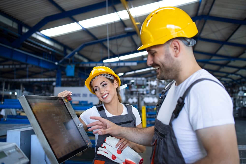 Zwei Arbeiter mit Schutzhelmen schauen in einer Fabrik auf einen Monitor.
